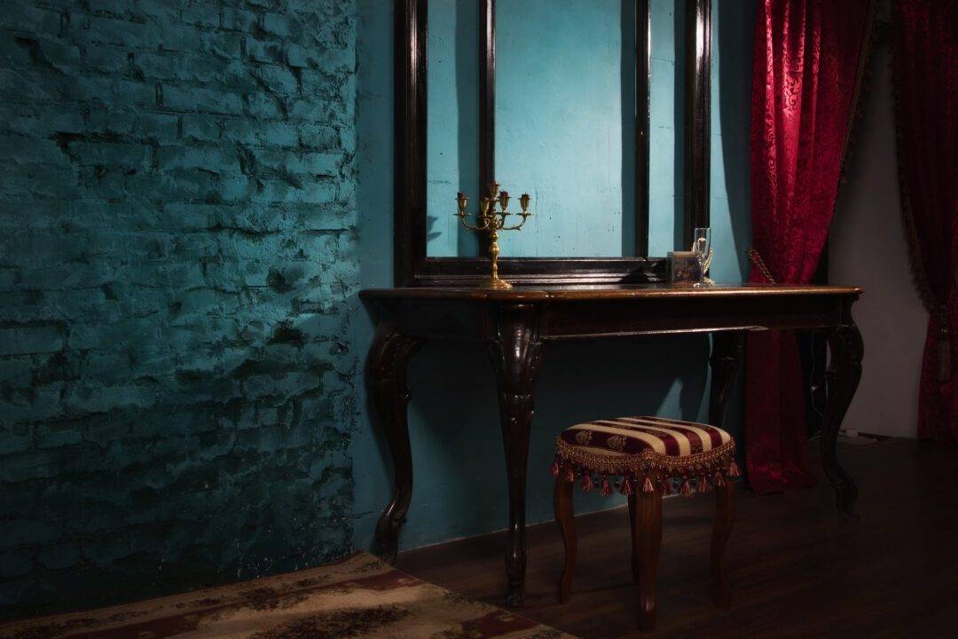 An elegant vintage-style vanity setup in a dimly lit room. The vanity table, with dark wood legs and a polished surface, holds a gold candelabra and a decorative box. Behind it, a large mirror with a black frame reflects the teal-painted brick wall. A small cushioned stool with red and beige striped upholstery and tassels is positioned in front of the vanity. Red curtains with a rich texture hang to the right, adding warmth to the space.