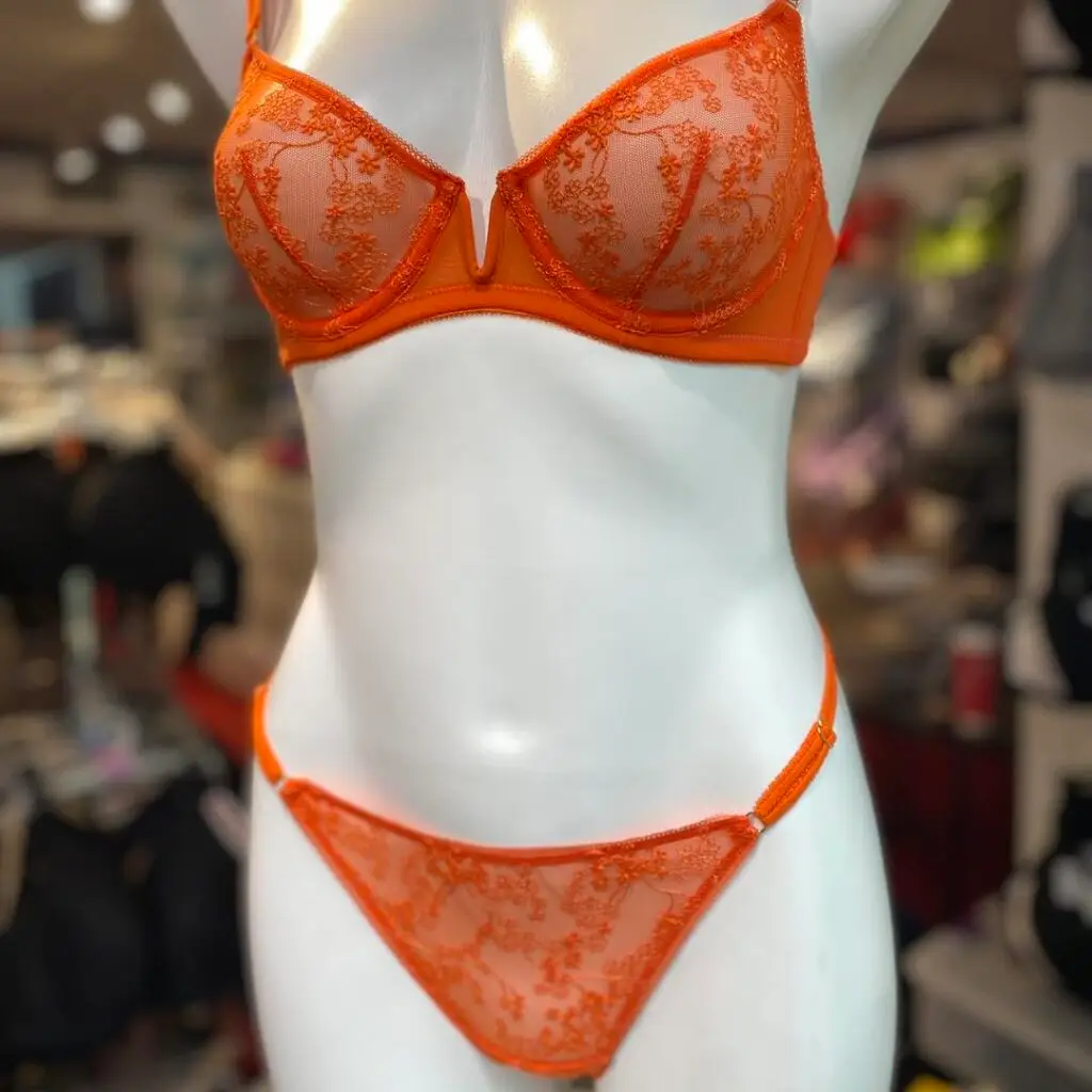 A vibrant orange lingerie set featuring a sheer lace bra with delicate floral embroidery and matching lace panties, displayed on a mannequin in a boutique setting."